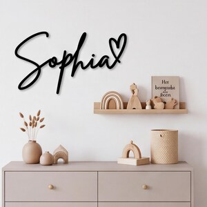 May include: A black wall-mounted sign featuring the name Sophia in a cursive script font with a heart symbol at the end. The sign is displayed above a wooden dresser and a floating shelf containing various wooden decorative items and a small book.