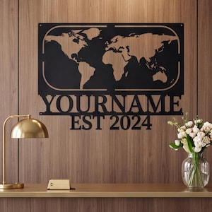May include: A black metal wall sign featuring a world map silhouette enclosed in a rectangular frame with rounded corners. Below the map, the text reads YOURNAME and EST 2024 in a bold, serif font. The sign is mounted on a vertical wood panel wall.