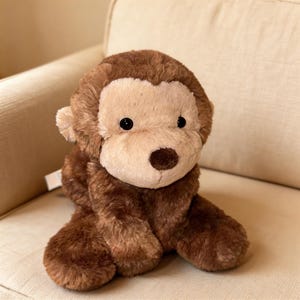 Könnte beinhalten: Ein Plüsch-Affe mit braunem und beigem Fell, schwarzen Augen und einer braunen Nase. Das Stofftier sitzt auf einem beigefarbenen Sofa. Der Affe hat einen freundlichen Ausdruck und besteht aus weichem Material.