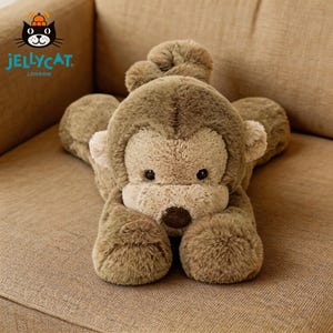 Puede incluir: Un peluche de mono en tonos marrones y beige, tumbado sobre una superficie de tono neutro. El juguete tiene una textura suave y peluda, con una cara clara y ojos oscuros. El logotipo de Jellycat London es visible.