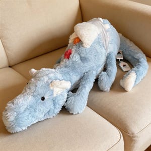 May include: A soft, pale blue dragon toy with white detailing on its head, wings, and tail. The dragon has a long body and tail, with orange and red patches. The toy is resting on a beige sofa.