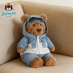 May include: A brown teddy bear dressed in a light blue and white jacket, trousers, and a hood with goggles. The bear is seated on a beige sofa. The jacket features a small orange graphic. The Jellycat London logo is in the top left corner.