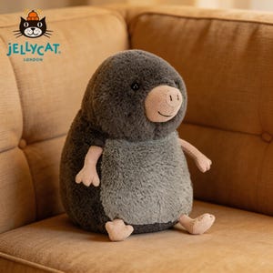 May include: A soft toy mole with a dark grey head and body, light grey belly, and pink nose, hands, and feet. The Jellycat London logo is visible. The plush animal is sitting on a brown sofa.