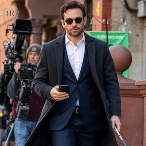 May include: A man in a dark gray overcoat, navy suit, and white shirt, wearing sunglasses, holding a smartphone. The man is walking on a city street, with a film crew in the background.