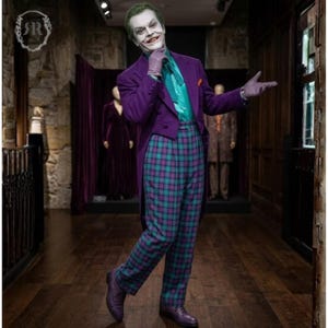 Puede incluir: Una figura vestida como el Joker, con un frac morado, una camisa verde azulado y pantalones a cuadros en tonos verde azulado y morado. La figura sonríe y posa en una habitación con pisos de madera oscura y paredes de piedra.