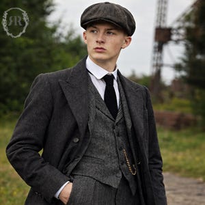 Puede incluir: Un traje de tres piezas gris carbón con una gorra plana a juego. El traje incluye una chaqueta cruzada, un chaleco y un pantalón. Se lleva debajo una camisa blanca con cuello y una corbata negra. Se ve una cadena dorada de reloj de bolsillo.