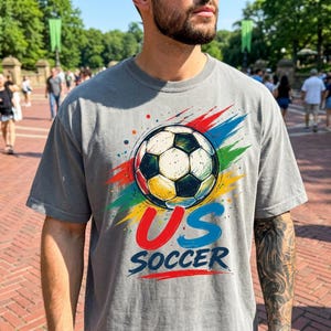 Op de afbeelding: Een grijs t-shirt met korte mouwen en een centrale afbeelding van een zwart-witte voetbal omringd door levendige rode, groene, gele en blauwe verfspatten. Onder de bal staat de tekst US SOCCER in dikke rode en blauwe letters gedrukt.