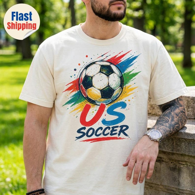 Op de afbeelding: Een cr&egrave;mekleurig T-shirt met korte mouwen en een afbeelding van een voetbal, gecentreerd boven de tekst US SOCCER. Het ontwerp bevat kleurrijke, verfachtige rode, blauwe en groene penseelstreken en spetters rondom de voetbalafbeelding.