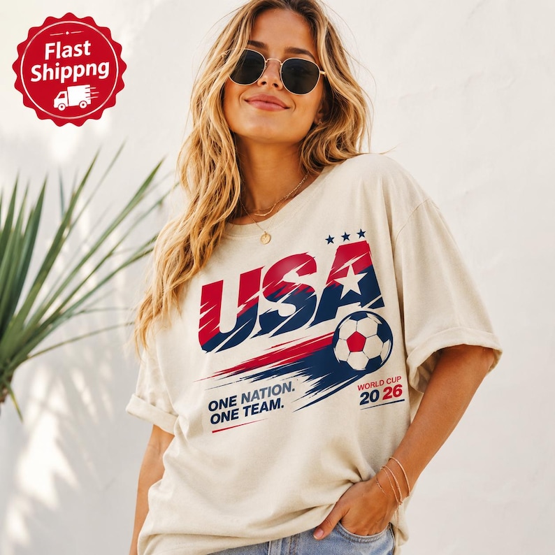 May include: A beige short-sleeve t-shirt featuring a graphic design with the text USA in red and blue, a soccer ball illustration, and the text ONE NATION. ONE TEAM. WORLD CUP 20 26. The design includes red and blue brushstroke accents.
