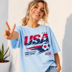 May include: A light blue t-shirt featuring a graphic with the text USA, ONE NATION. ONE TEAM., and WORLD CUP 20 26. The design includes a soccer ball, three stars, and red and dark blue brushstroke accents across the chest of the garment.