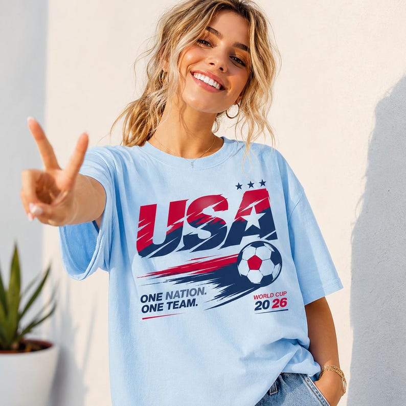 May include: A light blue t-shirt featuring a graphic with the text USA, ONE NATION. ONE TEAM., and WORLD CUP 20 26. The design includes a soccer ball, three stars, and red and dark blue brushstroke accents across the chest of the garment.
