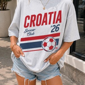 Pu&ograve; includere: Una t-shirt a maniche corte grigio chiaro con la scritta rossa CROATIA, la scritta blu Soccer Club, il numero 26 e una grafica di un pallone da calcio sopra strisce orizzontali rosse e blu. La maglietta &egrave; abbinata a pantaloncini di jeans azzurri.