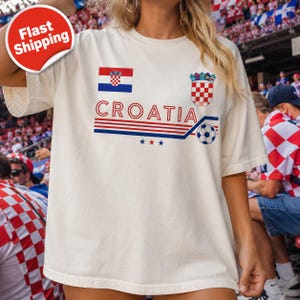 May include: An off-white short-sleeve t-shirt featuring the text CROATIA in red letters, the Croatian flag, the national coat of arms, a soccer ball graphic, and three small stars in red, white, and blue, with horizontal stripes across the chest.