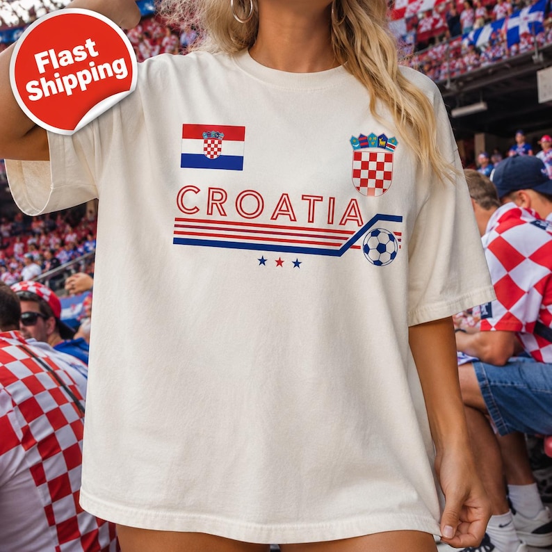 May include: An off-white short-sleeve t-shirt featuring the text CROATIA in red letters, the Croatian flag, the national coat of arms, a soccer ball graphic, and three small stars in red, white, and blue, with horizontal stripes across the chest.