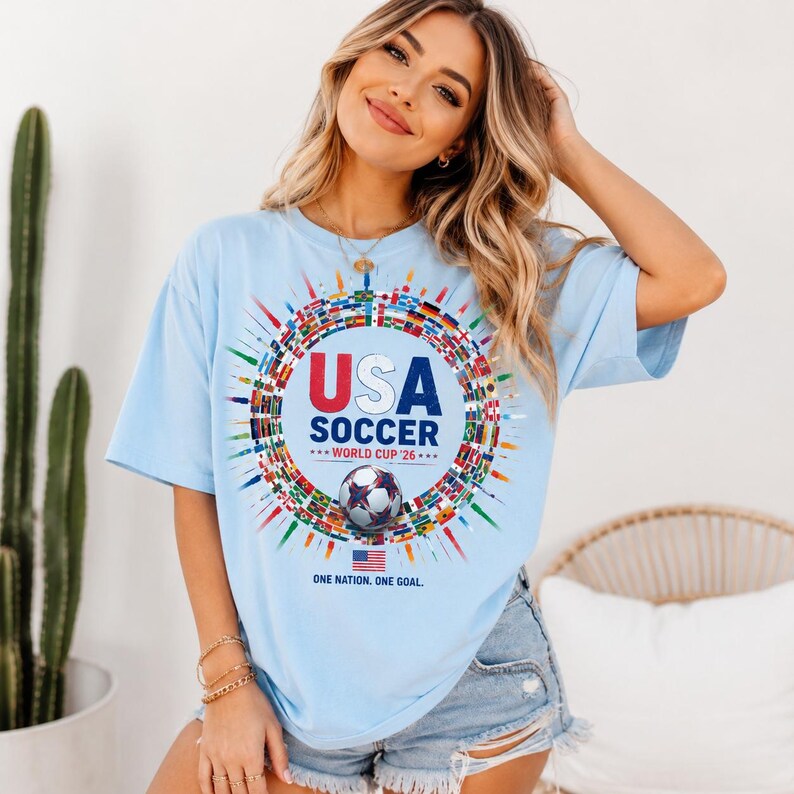 May include: A light blue t-shirt featuring a graphic of a soccer ball surrounded by a circular arrangement of international flags. The text reads USA SOCCER WORLD CUP '26, with the phrase ONE NATION. ONE GOAL. and a small American flag printed below.