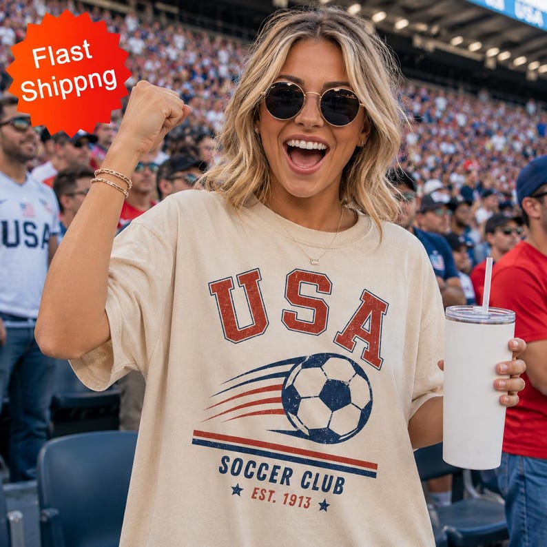 May include: A beige short-sleeved t-shirt featuring a graphic print with the text USA in red, a soccer ball with motion lines, and the text SOCCER CLUB EST. 1913 in blue. The shirt is worn by a person with blonde hair and sunglasses.