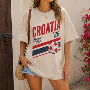 Pu&ograve; includere: Una t-shirt beige a maniche corte con la scritta rossa CROATIA, il numero 26 e le parole Soccer Club in blu. Una grafica di un pallone da calcio si trova sopra delle strisce orizzontali rosse e blu sulla parte anteriore del capo.