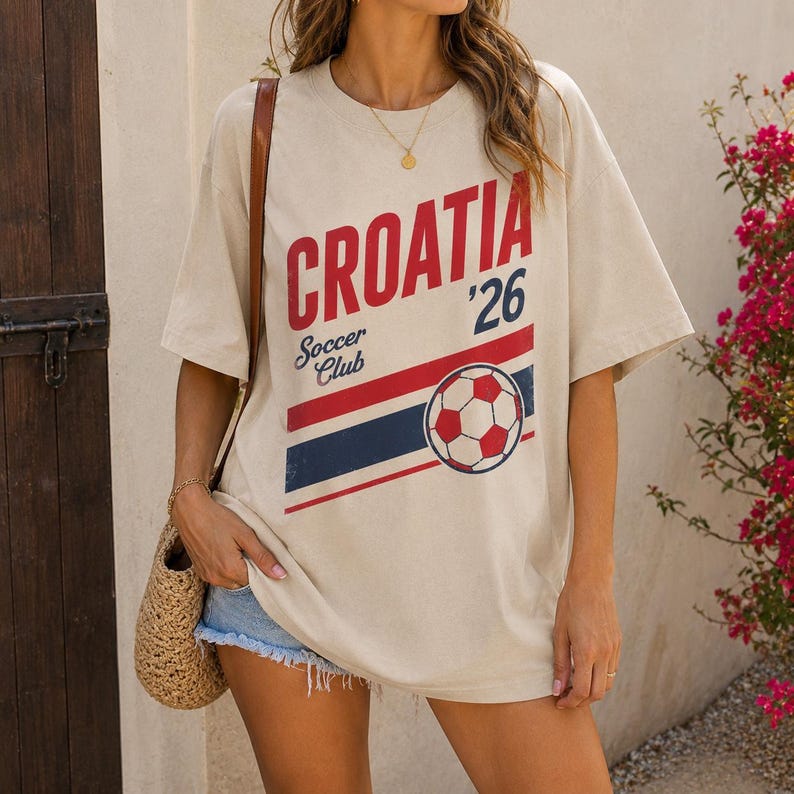 Pu&ograve; includere: Una t-shirt beige a maniche corte con la scritta rossa CROATIA, il numero 26 e le parole Soccer Club in blu. Una grafica di un pallone da calcio si trova sopra delle strisce orizzontali rosse e blu sulla parte anteriore del capo.