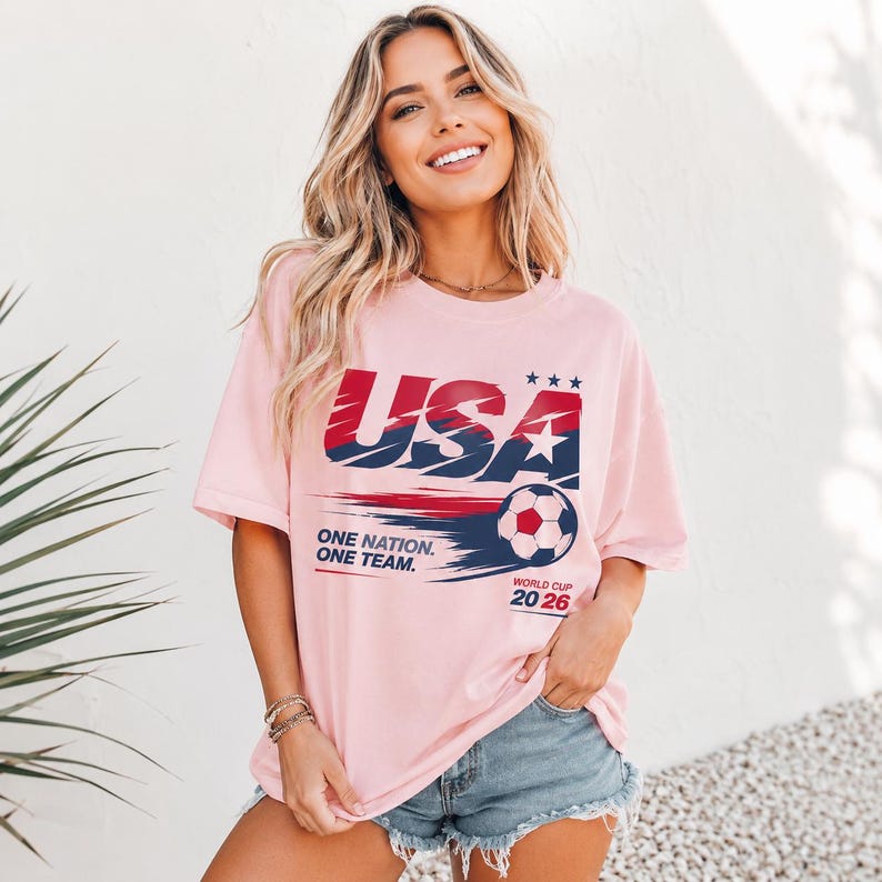 May include: A light pink short-sleeve t-shirt featuring a graphic with the text USA, ONE NATION. ONE TEAM., and WORLD CUP 2026. The design includes a soccer ball, three stars, and red and blue brushstroke accents across the front of the garment.
