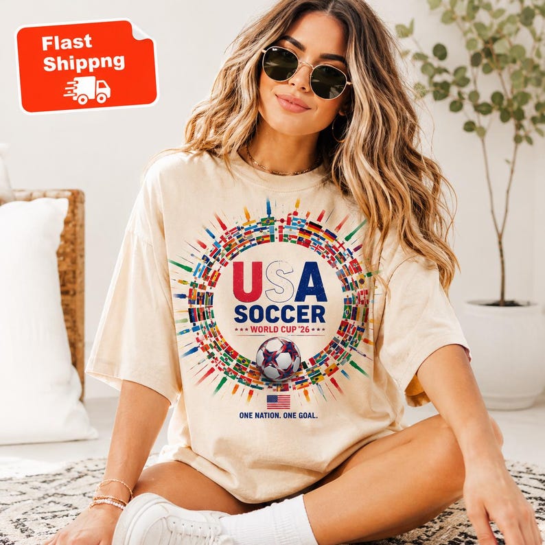 May include: A beige short-sleeve t-shirt featuring a graphic of a soccer ball surrounded by a circular arrangement of international flags. The text reads USA SOCCER WORLD CUP '26, ONE NATION. ONE GOAL. with a small American flag icon below the ball.