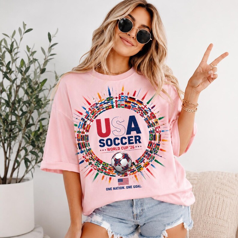 May include: A light pink short-sleeve t-shirt featuring a circular graphic of international flags surrounding the text USA SOCCER WORLD CUP '26, a soccer ball illustration, a small American flag, and the text ONE NATION. ONE GOAL.