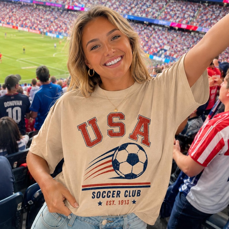 May include: A beige short-sleeve t-shirt featuring a graphic print with the text USA in red, a soccer ball with motion lines, and the text SOCCER CLUB EST. 1913 in blue and red. The shirt has a crew neckline and a relaxed fit design.