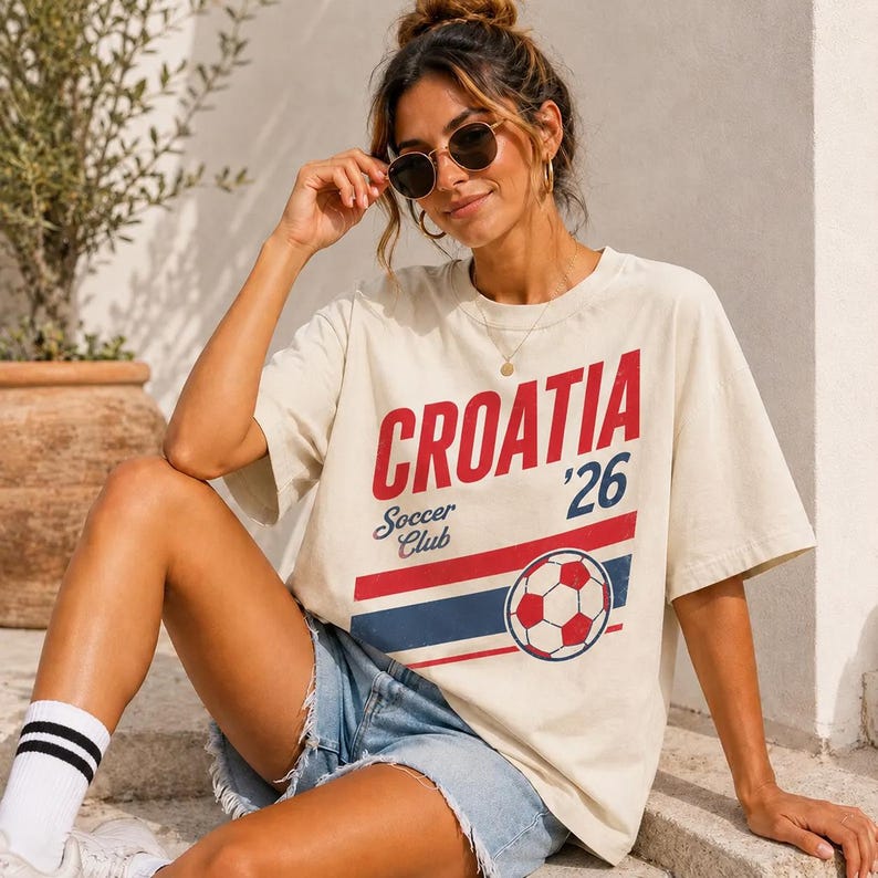 Pu&ograve; includere: Una t-shirt a maniche corte color bianco sporco con la scritta rossa CROATIA, la scritta blu '26 e la scritta blu Soccer Club. La maglia presenta una grafica di un pallone da calcio rosso e bianco sopra strisce orizzontali rosse e blu.