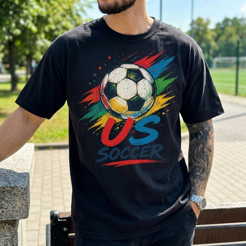 Op de afbeelding: Een zwart t-shirt met korte mouwen en een afbeelding van een voetbal boven de tekst US SOCCER. Het ontwerp bevat kleurrijke, abstracte penseelstreken in rood, geel, groen en blauw die achter de voetbalafbeelding uitstralen.
