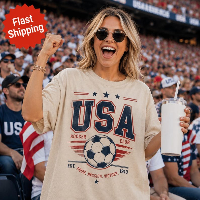 May include: A beige short-sleeved t-shirt featuring a graphic print with the text USA SOCCER CLUB EST. PRIDE. PASSION. VICTORY. 1913 and a soccer ball illustration. The design includes three stars above the text and horizontal lines.