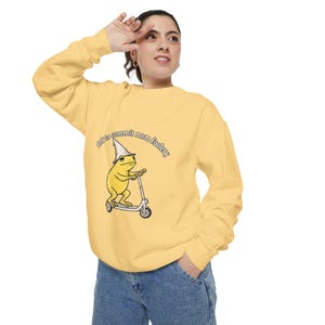 May include: A pale yellow long-sleeve sweatshirt featuring a graphic of a yellow frog wearing a pointed hat and riding a kick scooter. The text above the frog reads "Off to commit mom foolery" in a black, arched, serif font style.