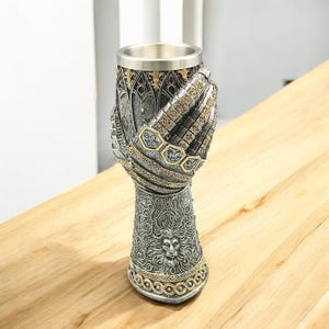 Puede incluir: Un cáliz decorativo con un diseño de guantelete de caballero medieval. El cáliz presenta un interior plateado y un exterior detallado con detalles dorados. La base del cáliz tiene un diseño de cabeza de león.