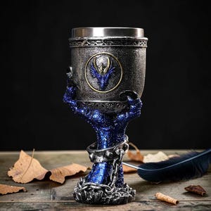 Puede incluir: Un cáliz decorativo con borde plateado y cuerpo gris oscuro texturizado. El cáliz está sostenido por una base de garra de dragón azul y presenta un emblema de dragón azul. La base está adornada con cadenas y detalles metálicos.