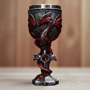 Peut inclure: Un gobelet décoratif avec un motif de dragon rouge. Le gobelet présente un bord et une base de couleur argentée, avec une sculpture de dragon détaillée en rouge et argent. Une gemme rouge est sertie au centre de la base.