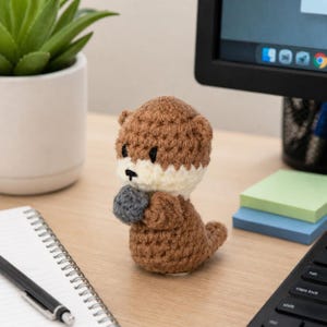 May include: A handmade crochet otter figurine. The otter has a brown head, body, and tail, with a cream-colored face and a gray ball-shaped hand. The otter is sitting on a wooden desk.