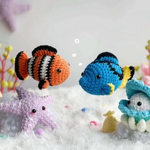 May include: Crocheted sea creatures including an orange and white striped clownfish, a blue fish, a lavender starfish, and a clam with a pearl. The handmade toys are set against a soft, white background.