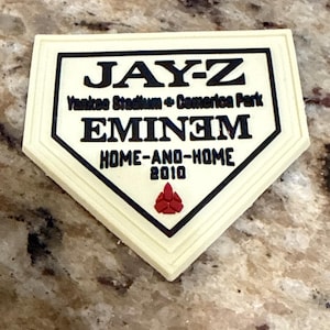 May include: A cream-colored, home plate-shaped commemorative item with black text. The text reads "JAY-Z," "EMINEM," "HOME-AND-HOME 2010," and "Yankee Stadium + Comerica Park." A red emblem is at the bottom.