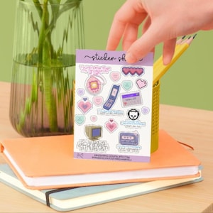 Puede incluir: Una mano sostiene una hoja de pegatinas con fondo morado. Las pegatinas presentan gráficos de temática retro, incluyendo un buscapersonas, un teléfono plegable y corazones con texto. Las pegatinas están sobre un cuaderno naranja.