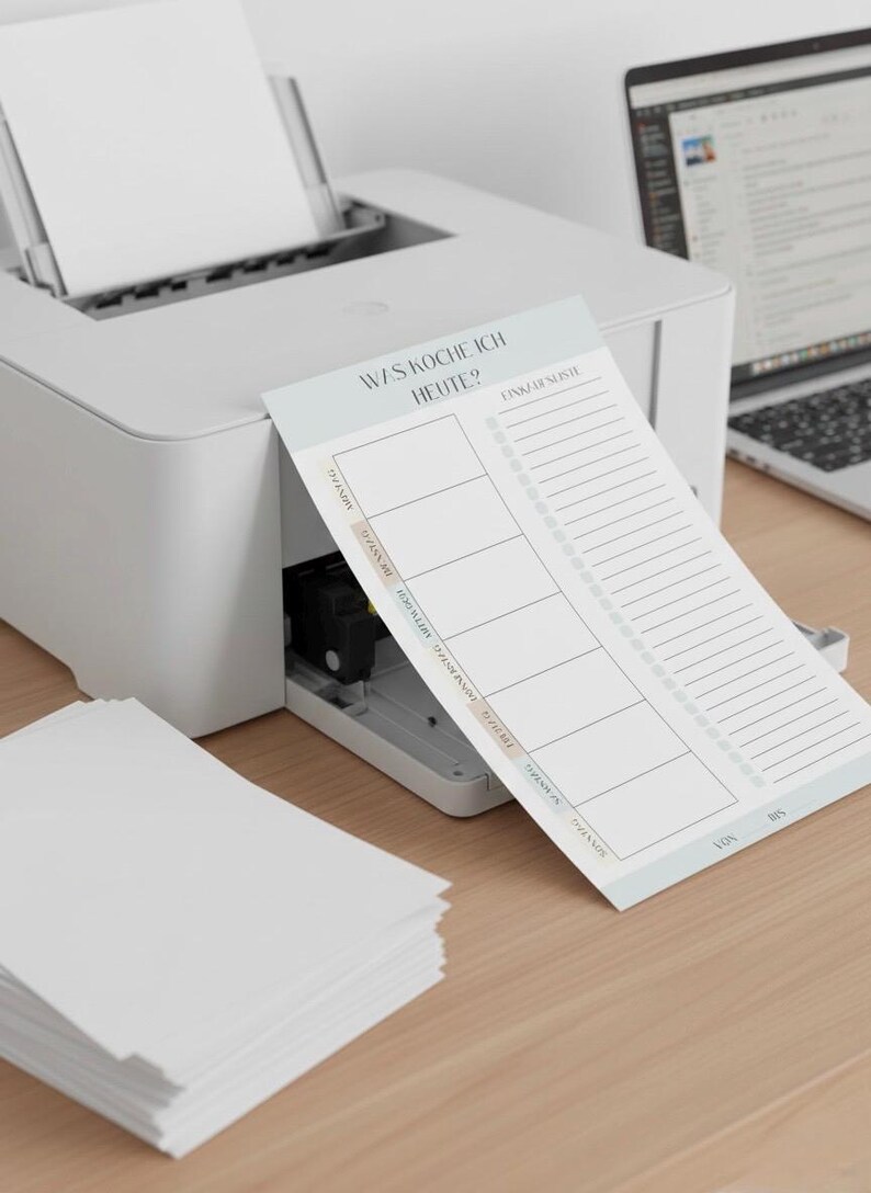 May include: A white printer next to a notepad with the text "WAS KOCHE ICH HEUTE?" and a shopping list. The notepad has sections for each day of the week. A stack of white paper is on the left.