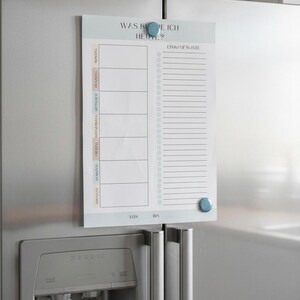 May include: A white magnetic weekly planner with the text "WAS KOE ICH HEUTE?" and "EINKAUFSLISTE" in German. The planner is attached to a stainless steel refrigerator with blue magnetic buttons. It includes sections for each day of the week and a shopping list.