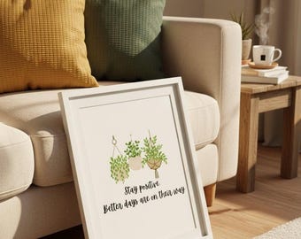 Op de afbeelding: Een ingelijste print met een botanische illustratie van hangplanten. Het kunstwerk bevat de tekst "Stay positive, Better days are on their way." De print wordt gepresenteerd op een beige bank met gele en groene kussens.