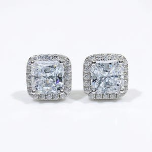 May include: A pair of square-cut, clear gemstone stud earrings. Each earring features a large, square center stone surrounded by a halo of smaller, clear gemstones. The earrings are set in a silver-colored metal.