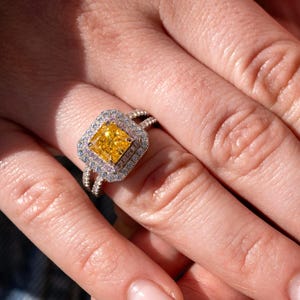May include: A square-cut yellow diamond ring with a double halo of smaller diamonds. The ring has a split shank band. The ring is set on a finger, with the skin tone in the background.