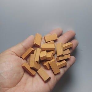 Puede incluir: Un puñado de pequeños ladrillos rectangulares de color marrón anaranjado. Los ladrillos son de tamaño y forma uniformes y parecen estar hechos de un material sólido. Los ladrillos se sostienen en una mano abierta sobre un fondo gris.