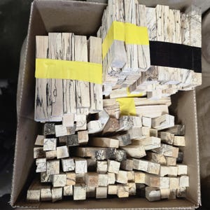 Könnte beinhalten: Ein Karton voller Holzblöcke in verschiedenen Größen und hellen Farben. Einige Blöcke sind mit gelbem Klebeband gebündelt, andere lose. Das Holz weist dunkle, komplizierte Maserungsmuster auf.