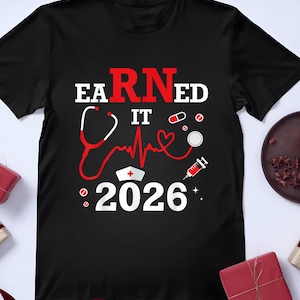 Könnte beinhalten: Schwarzes T-Shirt mit einem Design für Krankenschwestern. Das Design enthält die Worte "EARNED IT" in Weiß und Rot, ein Stethoskop, eine Herzschlaglinie, eine Krankenschwestermütze, Pillen und das Jahr "2026".