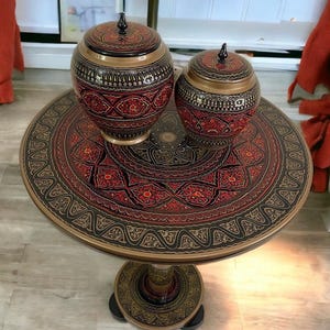 Op de afbeelding: Twee decoratieve houten potten met deksels staan op een ronde tafel. De potten en de tafel hebben ingewikkelde patronen in rood, zwart en goud. De tafel heeft een voetstuk en staat op een lichtgekleurde vloer.