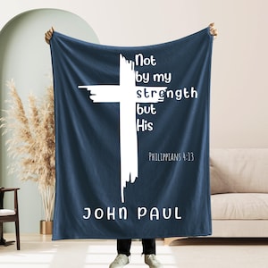 May include: A navy blue blanket with a large white cross design and the text "Not by my strength but His" and "Philippians 4:13" and "JOHN PAUL". The blanket is held up by a person.