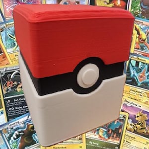 May include: A red, black, and white storage box, resembling a Poké Ball, is displayed. The box is rectangular with a circular button detail. Trading cards are visible in the background. The box is designed for storing collectible cards.