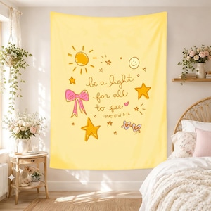 May include: A rectangular yellow wall tapestry featuring the hand-drawn text 'be a light for all to see -MATTHEW 5:16'. The design includes illustrations of a sun, stars, a pink bow, a smiley face, and heart-shaped sunglasses in orange and pink.