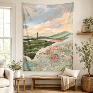 May include: A rectangular wall tapestry featuring a painted landscape of a grassy hill with a dark cross at the peak. The scene includes a foreground of wildflowers in shades of pink and green, set against a soft, cloudy sky in peach and blue tones.